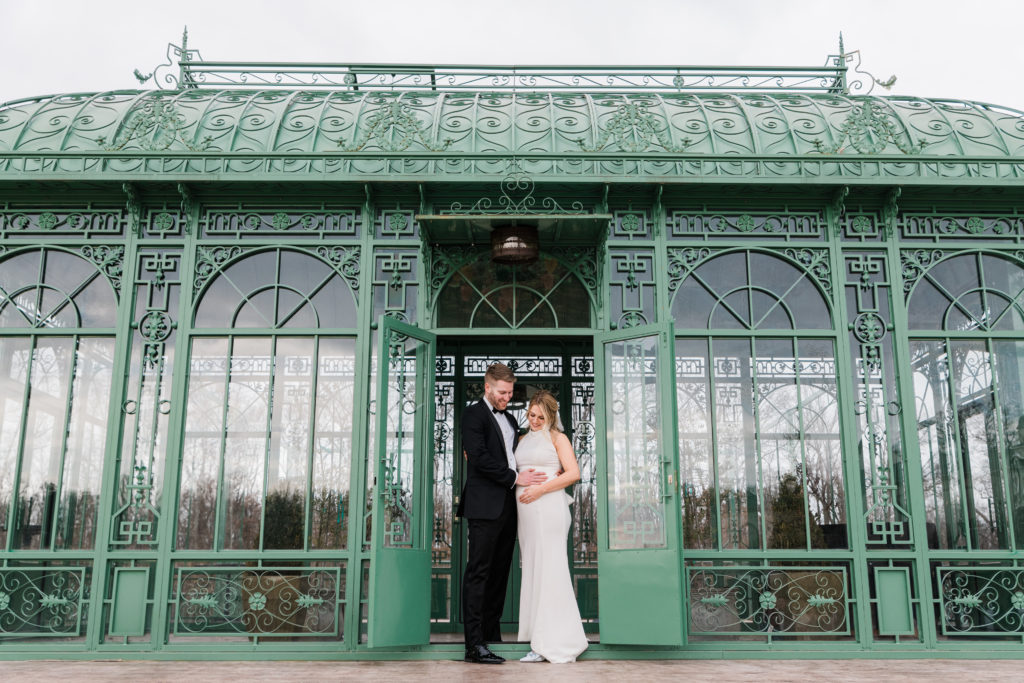 A bride and groom posing together in front of a green greenhouse at Morais Vineyards and Winery