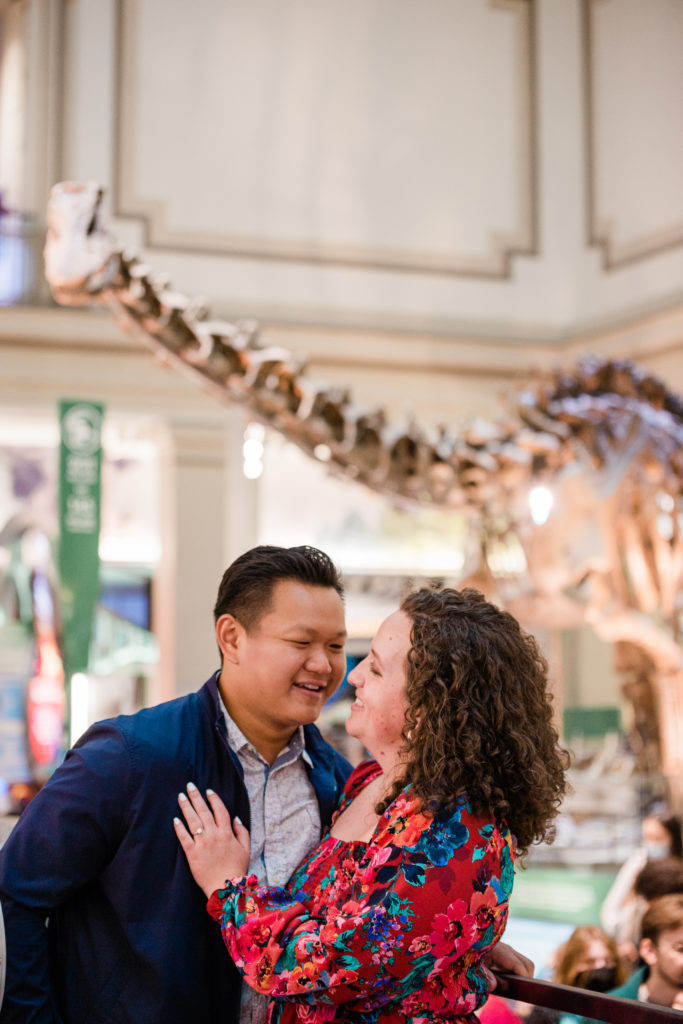 A man and a woman hugging with a dinosaur fossil in the background at the Smithsonian Natural History Museum