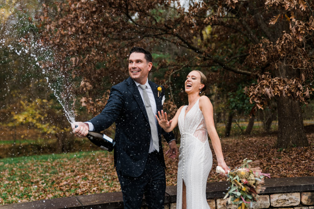 A groom pops champagne while standing next to a bride