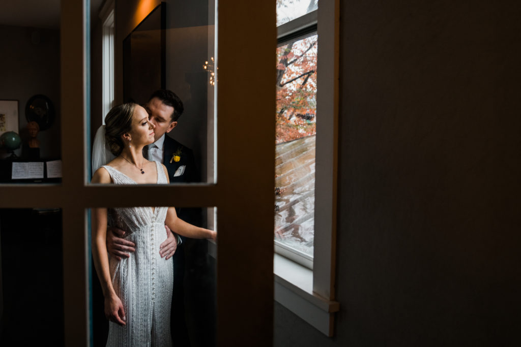 A bride and groom pose by a window