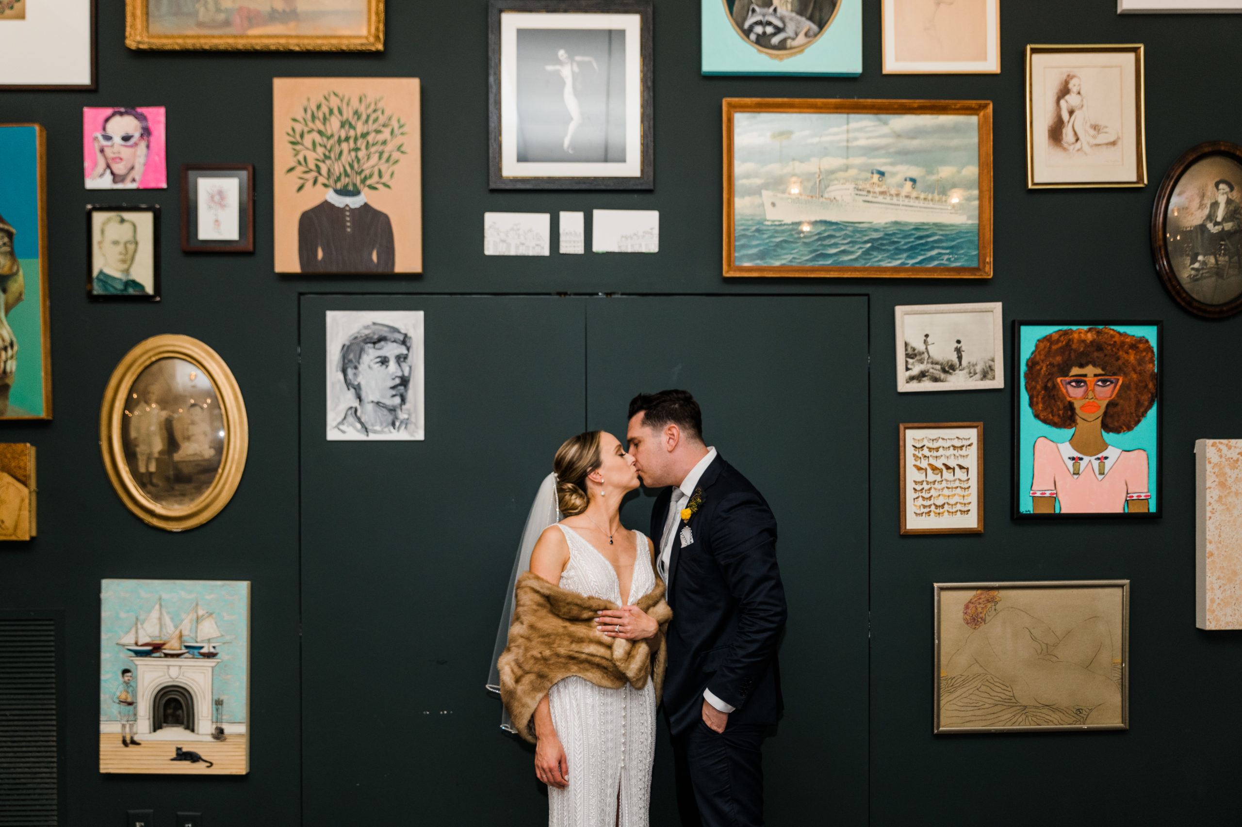 A bride and groom kiss against a blue wall with lots of pictures on it