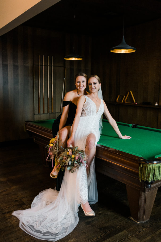 Bride poses on top of a pool table with a bridesmaid
