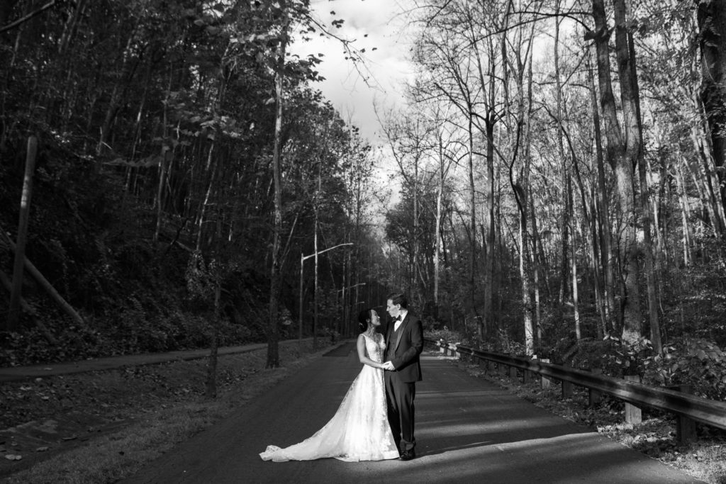 Black and white picture of a bride and groom embracing on a road surrounded by trees