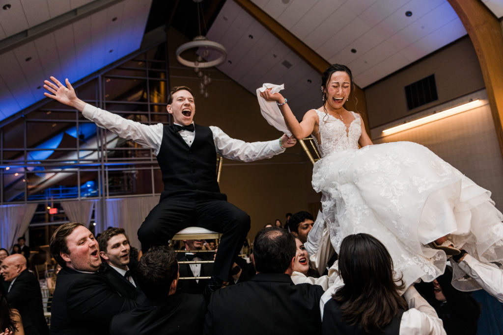 A bride and groom sitting in chairs and being raise up by a crowd