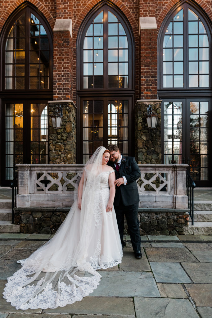 A bride and groom pose outside in front of a building with tall windows