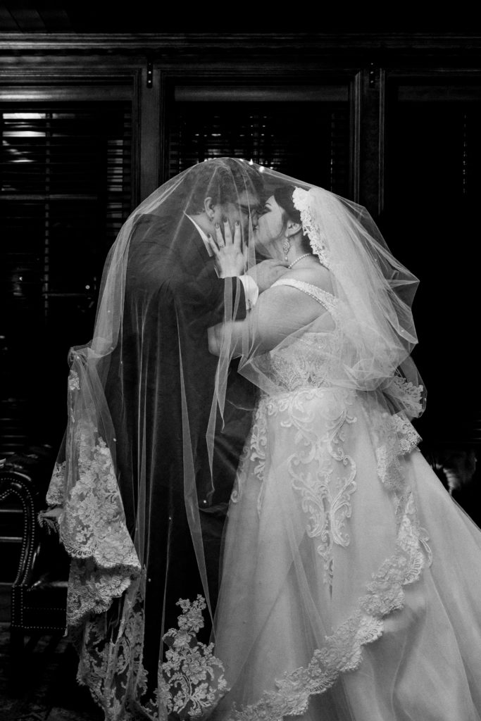 A black and white picture of a bride and groom kissing under a lace veil