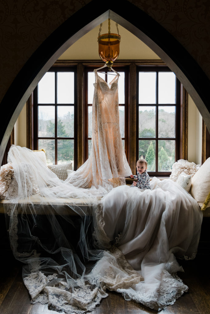 A white wedding dress hanging in a window and a baby sitting on the skirt