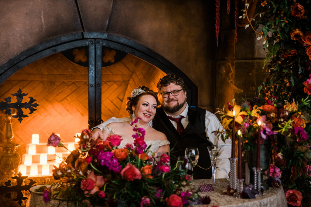 A bride and groom sitting together in front of a fireplace with colorful flowers in front of them