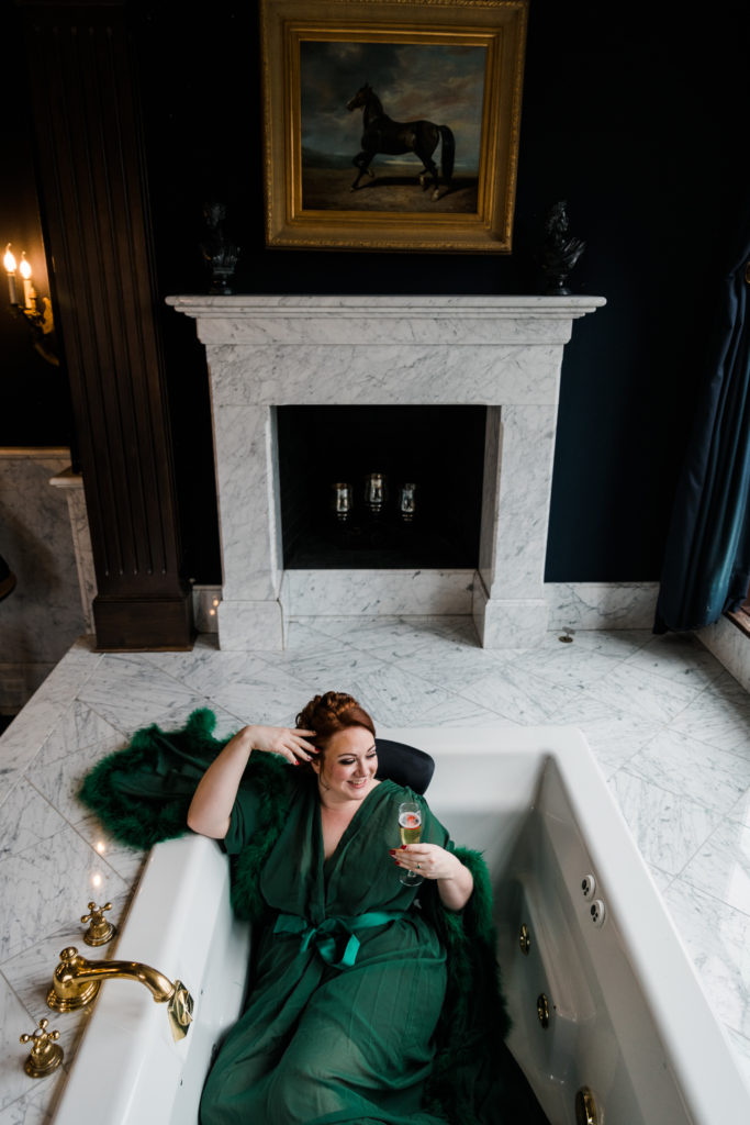 A woman in a green robe sits in a marble bath tub with a glass of champagne