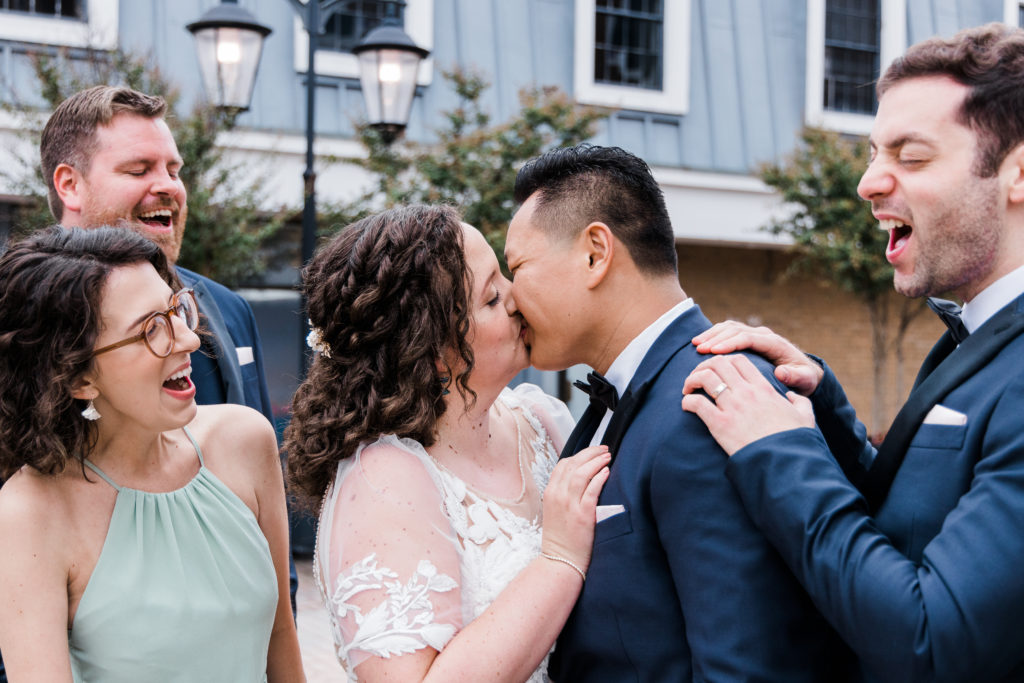 A bride and groom kiss with people standing around them cheering