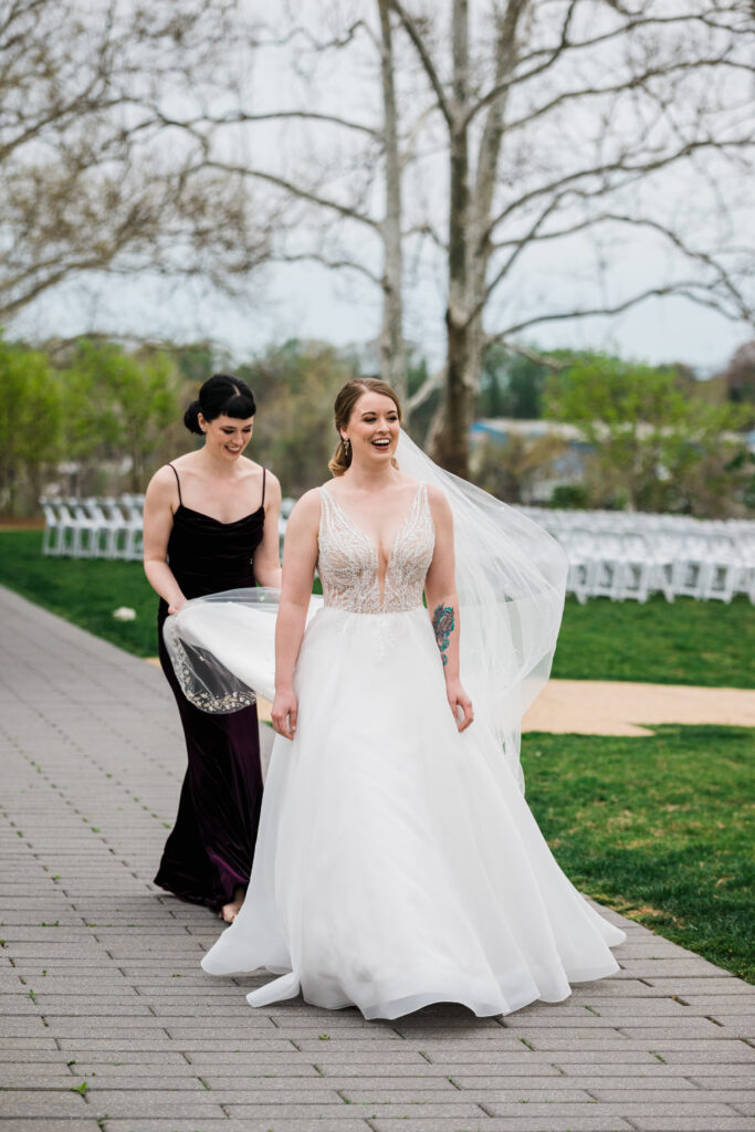 A woman in a wedding dress walking while a woman behind her holds the back of her dress
