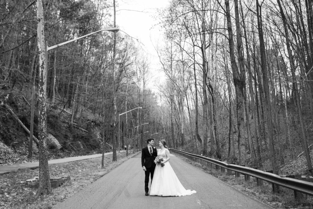 A black and whit picture of a man in a suit and a woman in a wedding dress standing on a road surrounded by trees