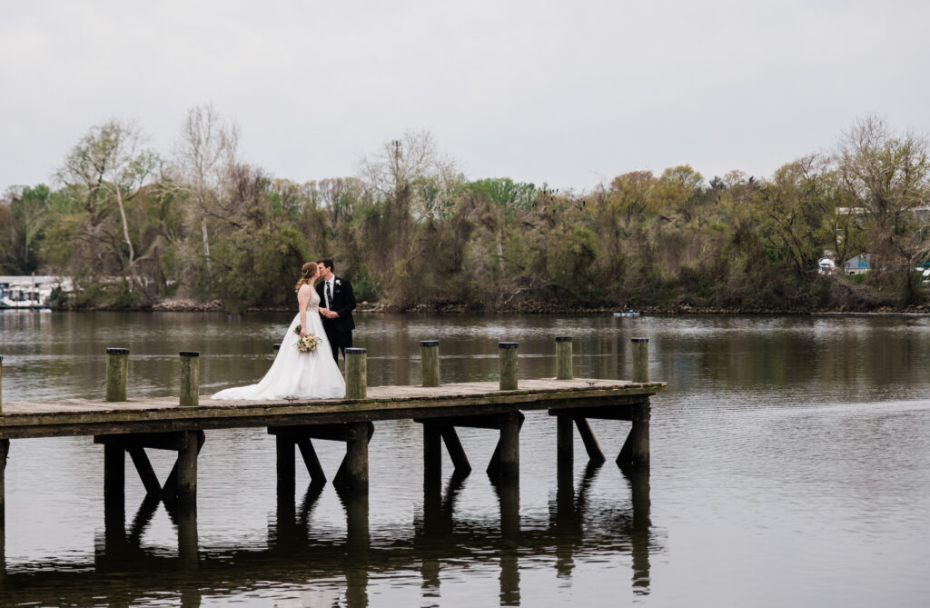 A man in a suit and a woman in a wedding dress standing on a dock kissing