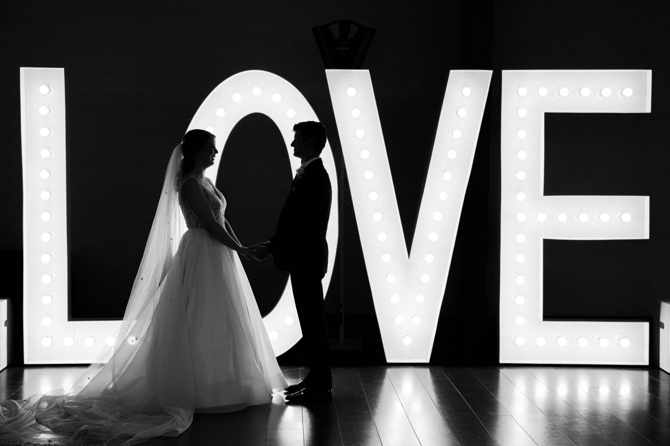A couple holding hands in front of a large, lit up sign that says "LOVE"