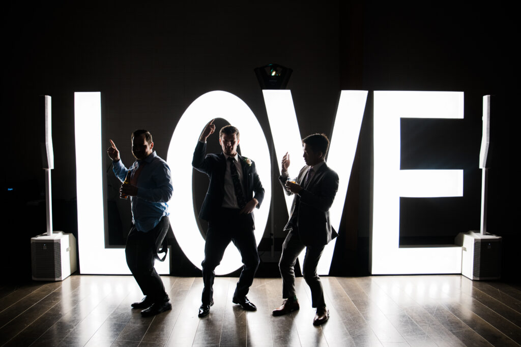 3 men standing in front f a large lit up sign that says Love