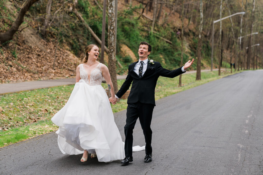 A man in a suit and a woman in a wedding dress walking down a road holding hands and laughing