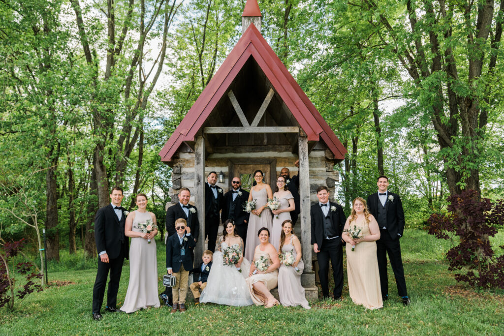 A group of people dressed up in dresses and suits standing in front of a small log cabin