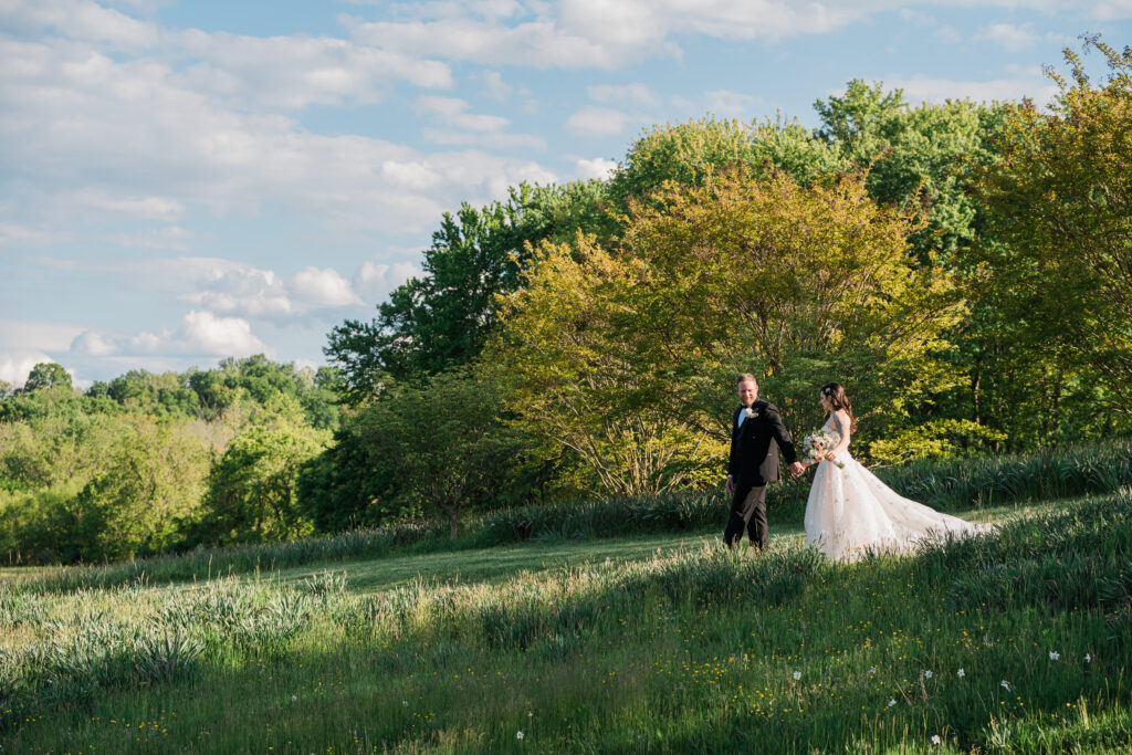 A bride and groom walking hand in hand in a field with trees