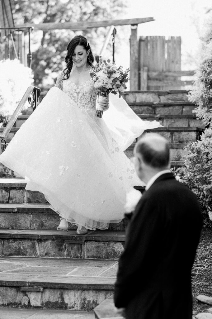 A woman in a wedding dress holding flowers and walking down a staircase towards a man waiting at the bottom of the stairs