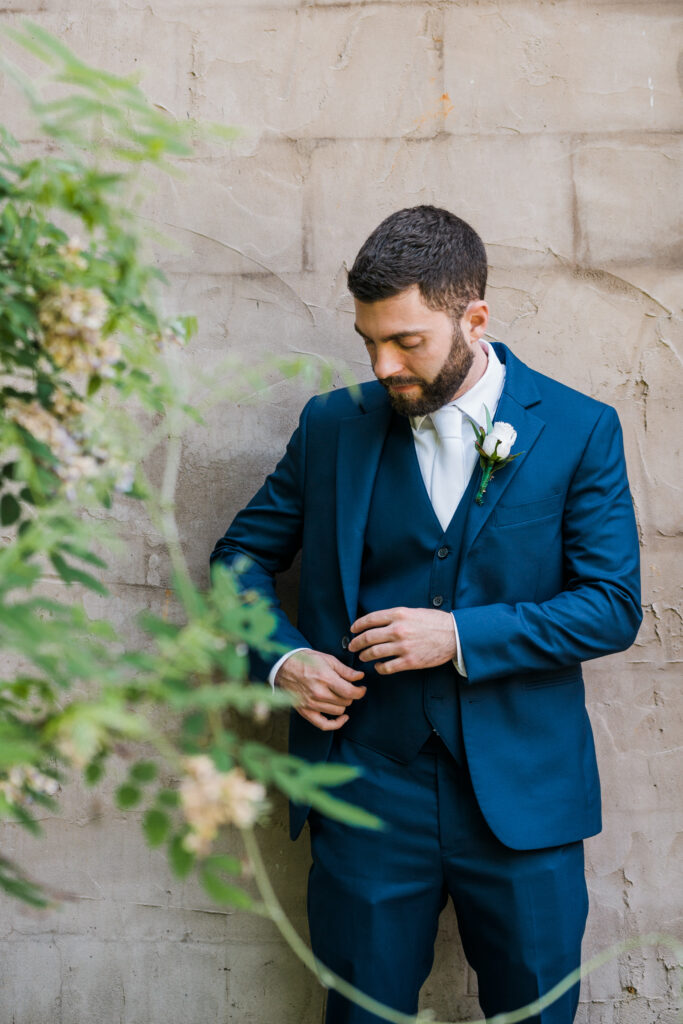 A groom standing next to a wall while fixing his jacket.